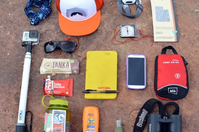 Day-hike essentials laid out on red ground: hat, sunscreen, water bottle, snacks, phone, map, and first-aid kit.