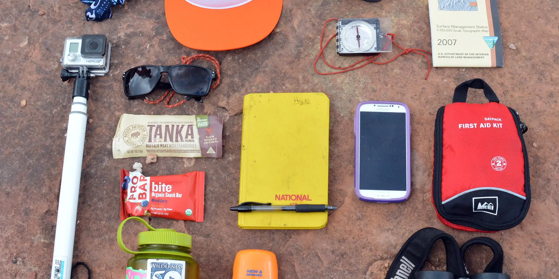 Day-hike essentials laid out on red ground: hat, sunscreen, water bottle, snacks, phone, map, and first-aid kit.