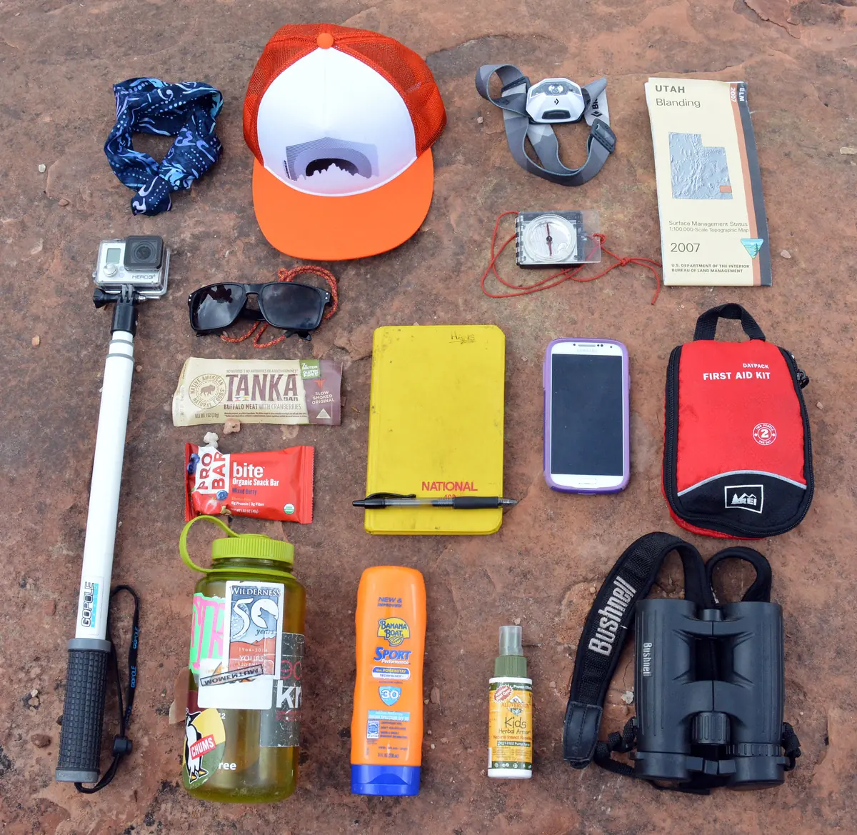 Day-hike essentials laid out on red ground: hat, sunscreen, water bottle, snacks, phone, map, and first-aid kit.