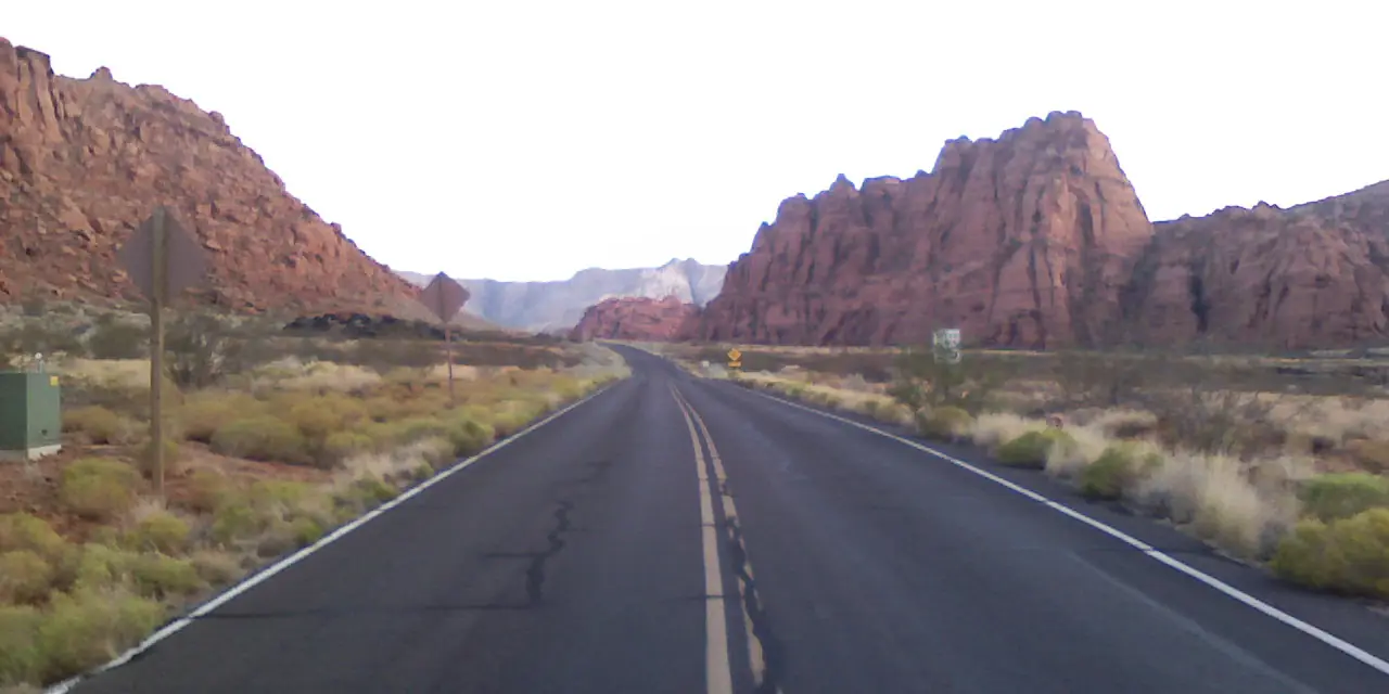 A two-lane road leading toward red and white sandstone cliffs at the entrance to Snow Canyon.